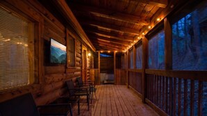 Una cabaña de madera con porche, sillas, un televisor y vistas al bosque.