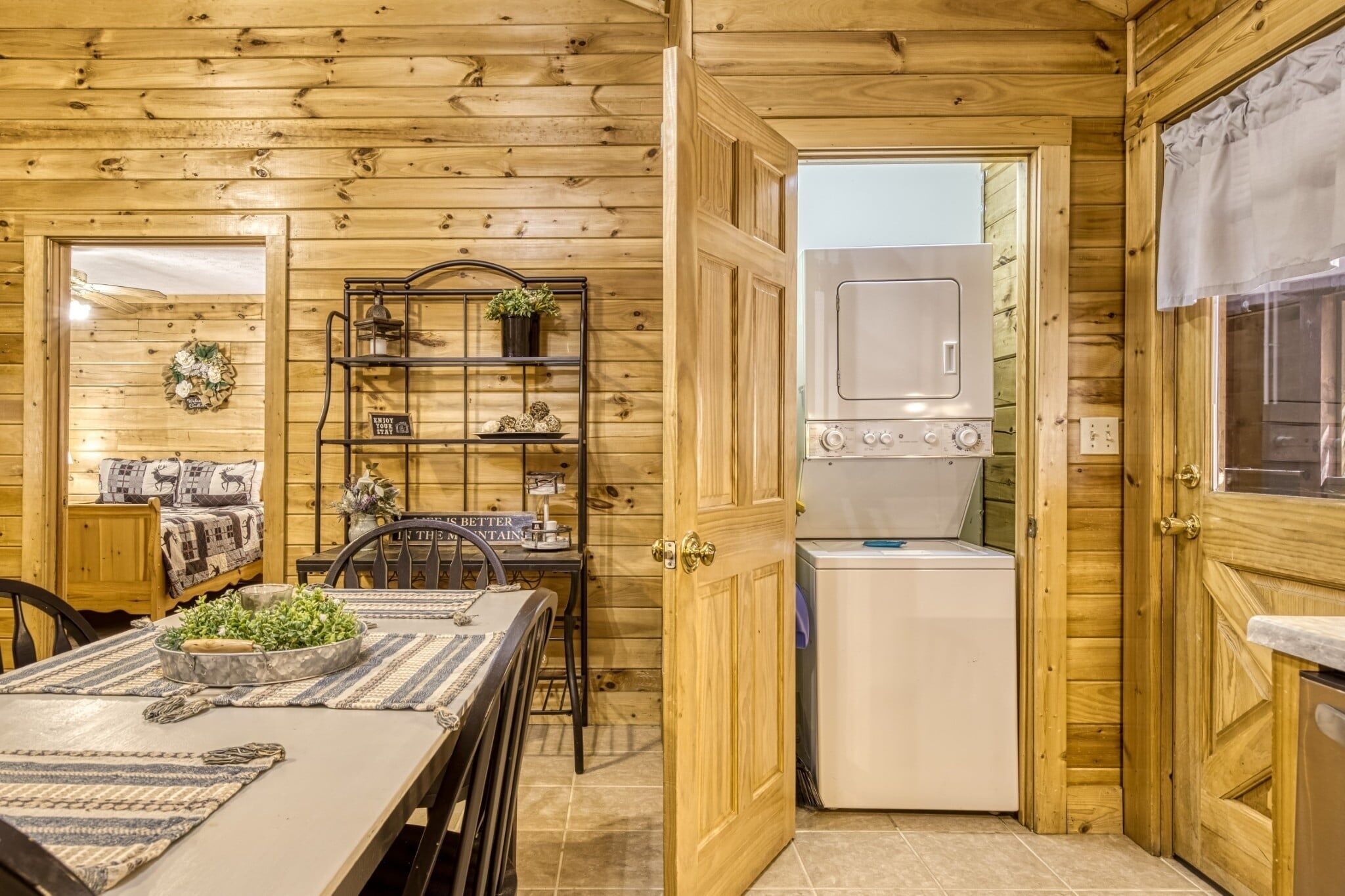 A wooden interior with a dining table, chairs, a shelf with decorative items, a washing machine, and a door leading to another room.