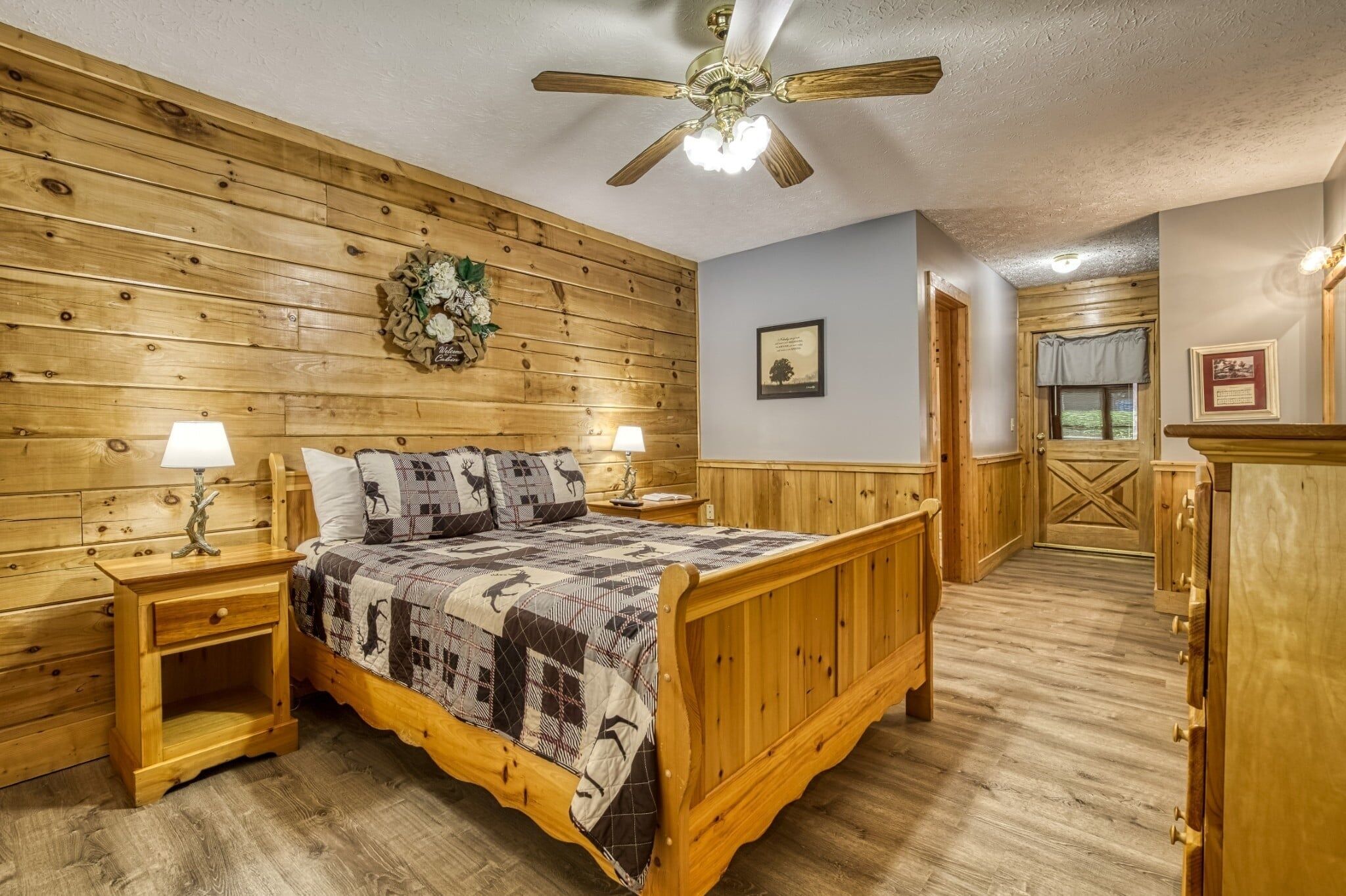 A wooden-paneled bedroom with a bed, nightstands, a ceiling fan, and a door leading to another room.