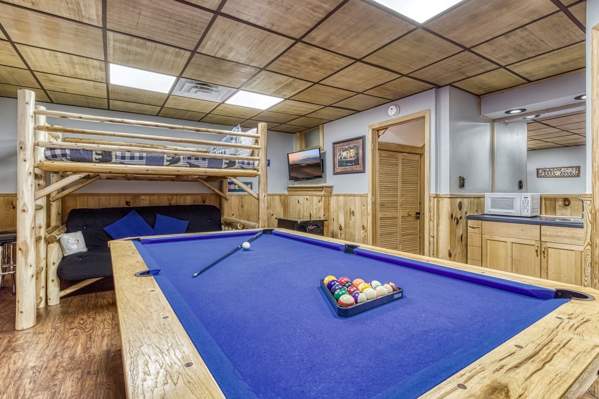 A pool table with a blue felt surface, a wooden bunk bed with a mattress and pillows, a television mounted on the wall, and a microwave on a wooden counter.