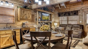 Interior rústico de una cabaña con una mesa de comedor de madera, sillas, chimenea y televisor.