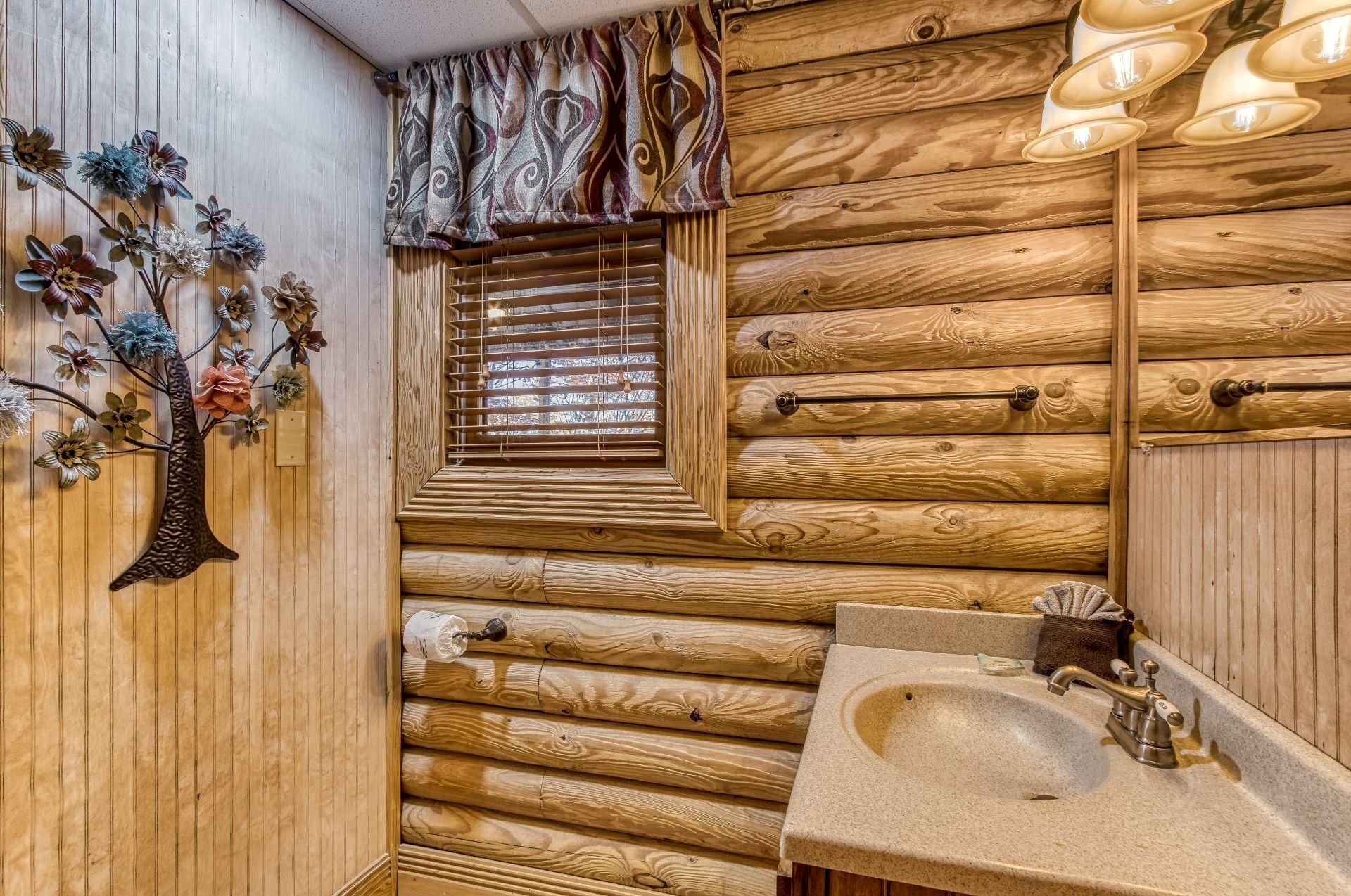 A rustic wooden cabin bathroom with a wooden slat window, a sink with a single faucet, and decorative flowers on the wall.