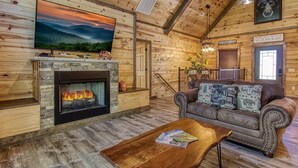 L’intérieur chaleureux d’un chalet avec une cheminée en pierre, un canapé en cuir et un téléviseur fixé au mur.