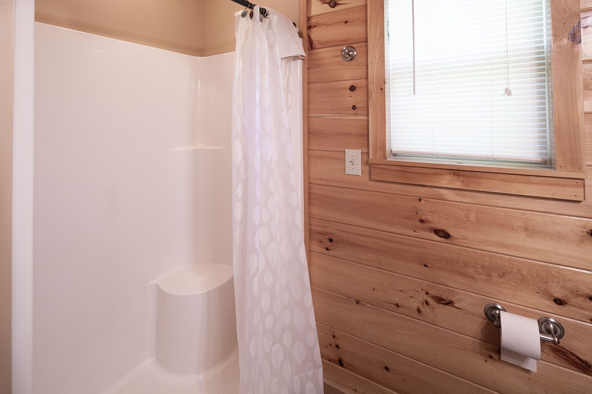 A wooden-paneled bathroom with a shower curtain, a toilet paper holder, and a window with blinds.
