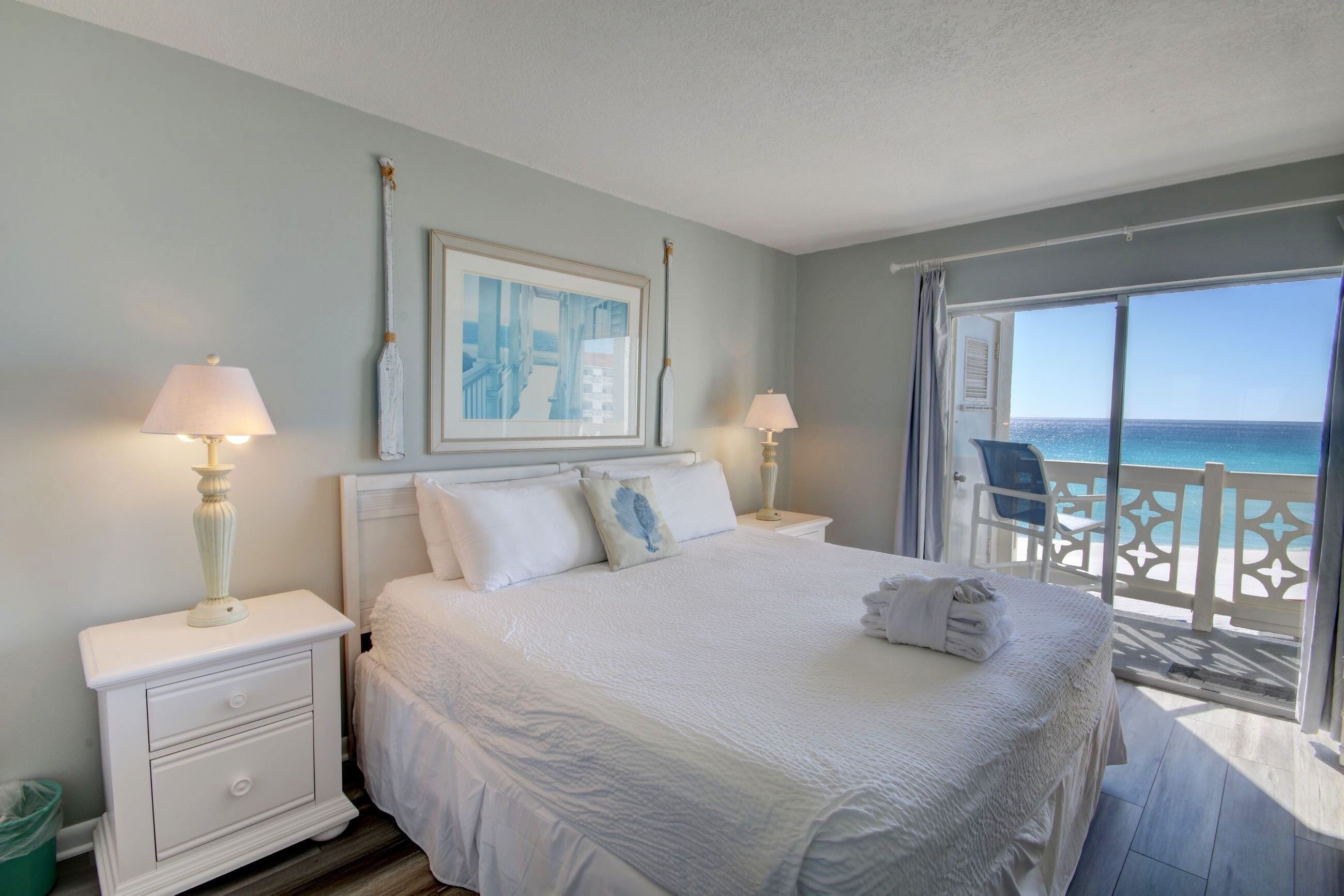A neatly made bed with white linens, a framed picture on the wall, and a view of the ocean through a balcony.