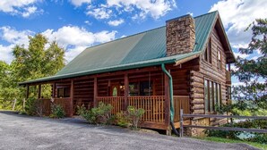 A log cabin with a green metal roof, a stone chimney, and a wooden porch.