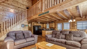 A wooden cabin interior with two sofas, a coffee table, a kitchen area, and a staircase.