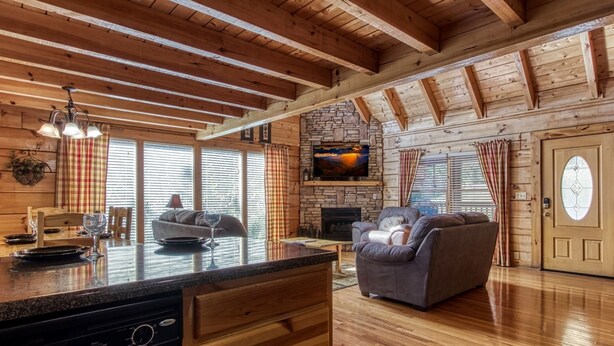 A cozy cabin interior with a stone fireplace, wooden ceiling beams, a dining area, and a living room with a sofa and a TV mounted on the wall.