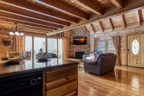 A cozy cabin interior with a stone fireplace, wooden ceiling beams, a dining area, and a living room with a sofa and a TV mounted on the wall.