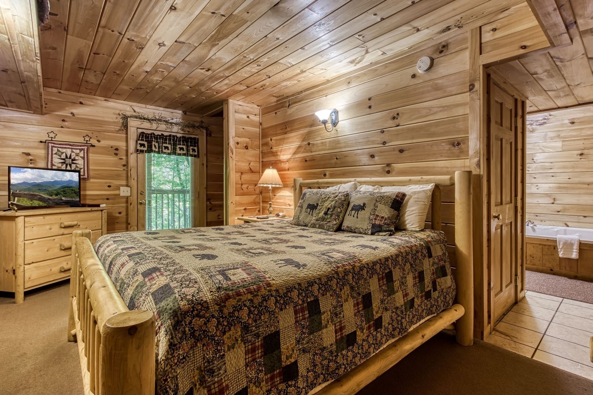 Een gezellig houten slaapkamer met een bed, nachtkastje, lamp, televisie en een raam met uitzicht op de natuur.