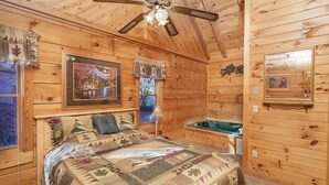 A cozy wooden cabin bedroom with a bed, a window with curtains, a framed painting, and a ceiling fan.