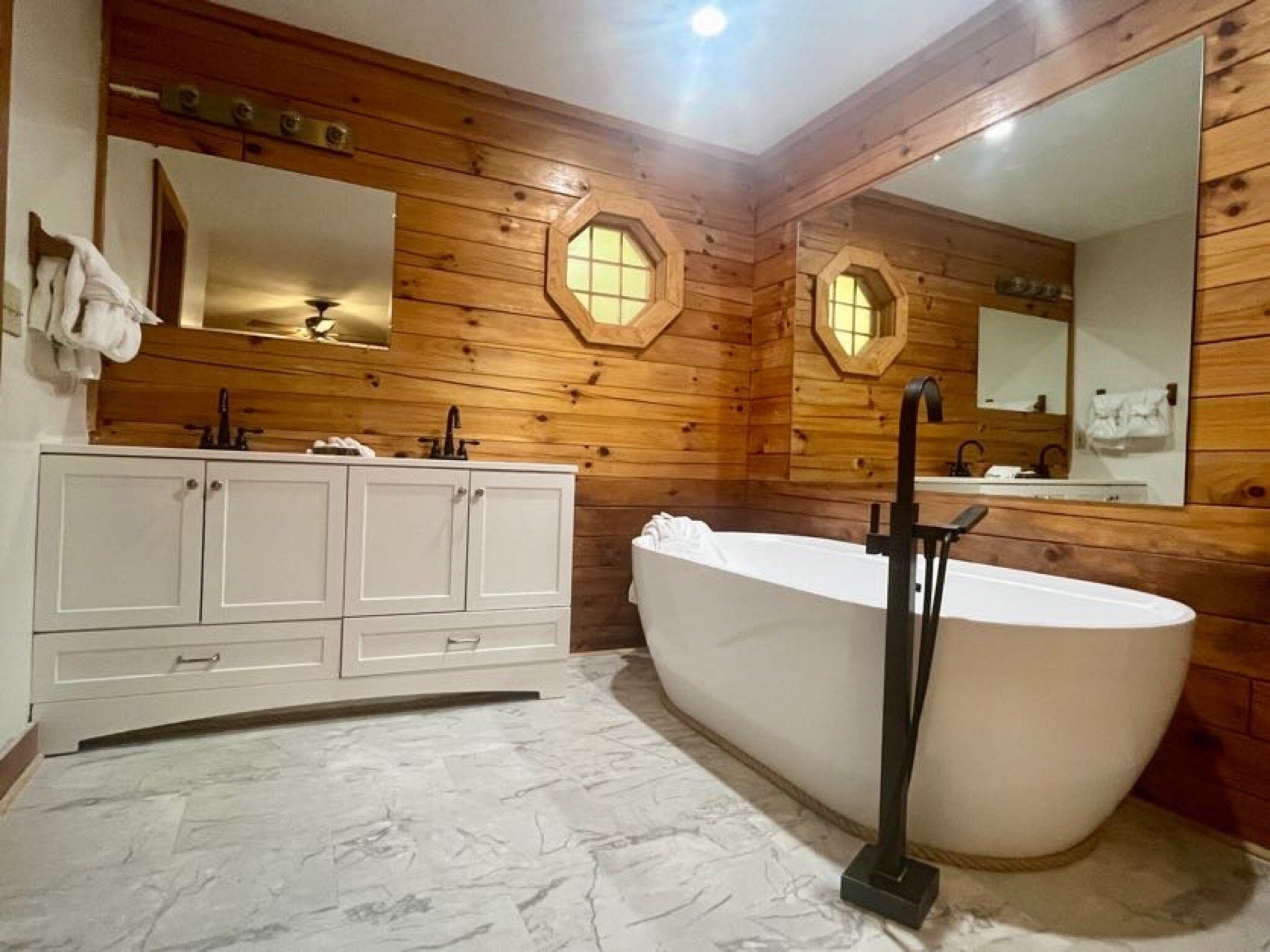 Un baño de estilo cabaña de madera con una bañera independiente, un tocador con espejo y suelo de mármol.
