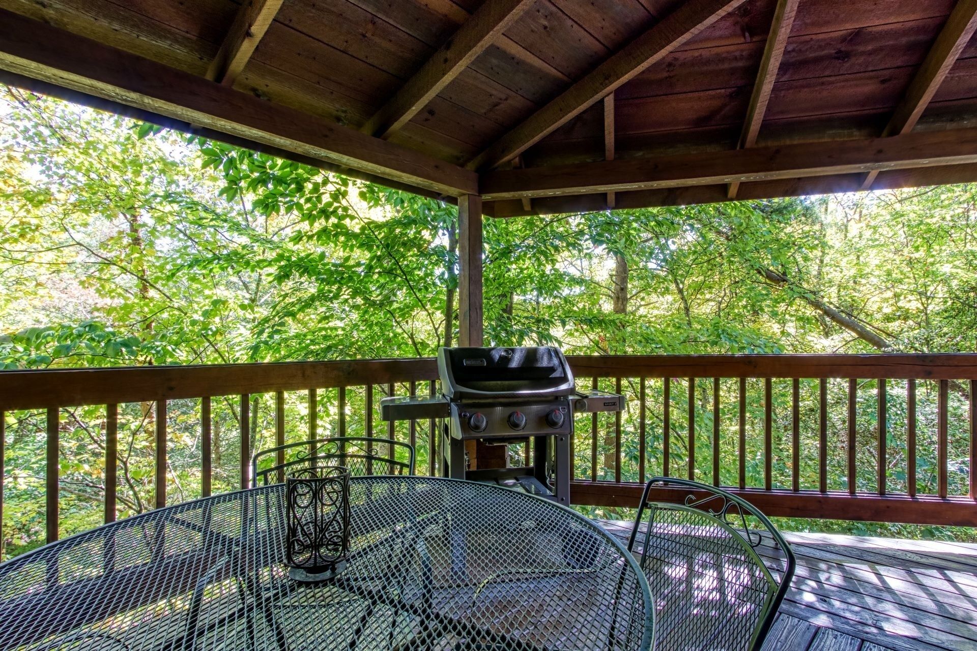 A covered outdoor area with a metal table and chairs, a grill, and a view of a wooded area.