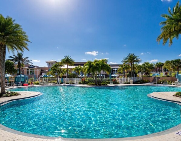 A clear blue swimming pool surrounded by palm trees and a building with a red roof in the background.