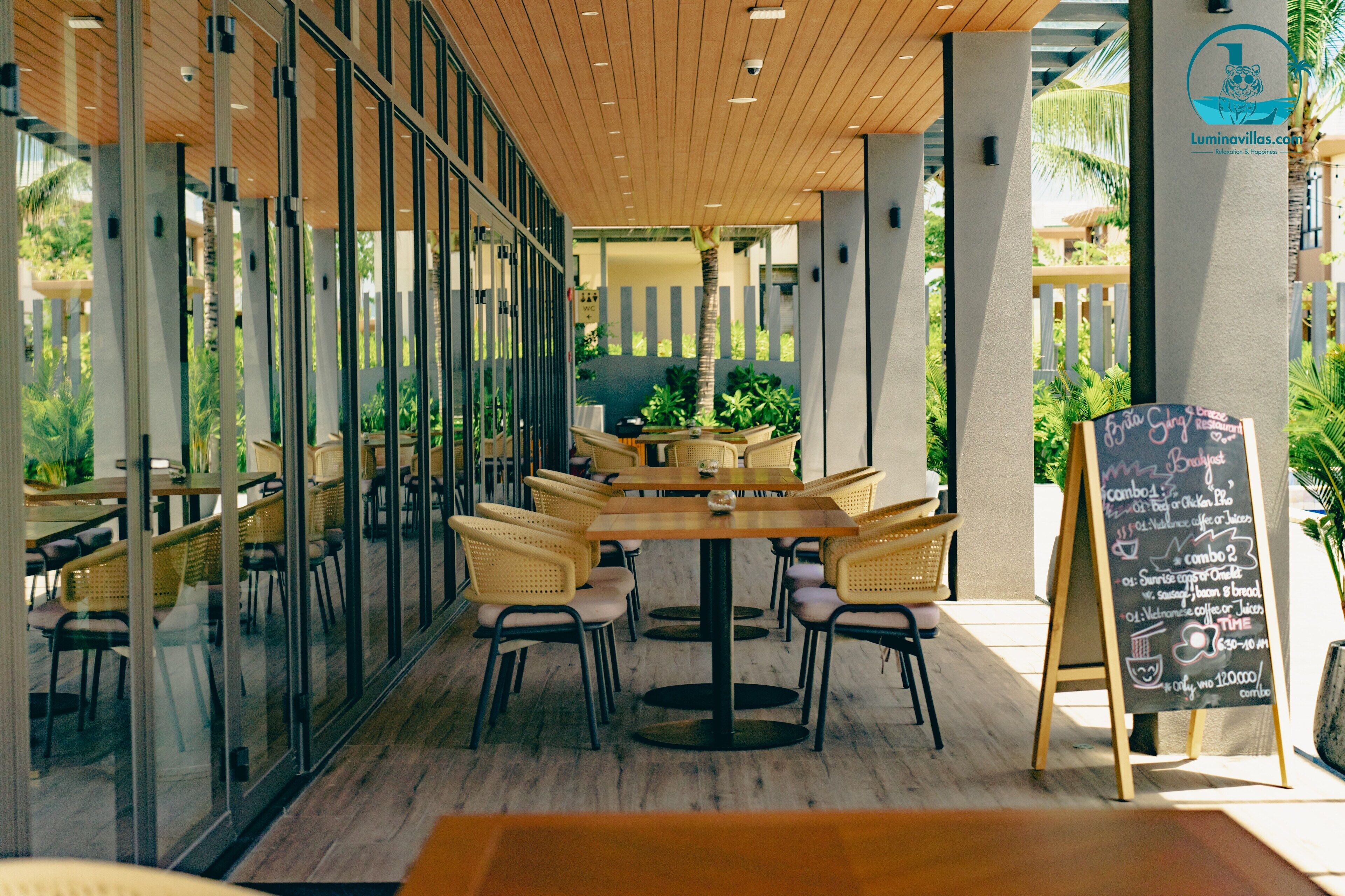 Un espace de restauration en plein air avec des tables et des chaises en bois, un tableau noir servant de menu et des murs en verre offrant une vue sur le jardin.