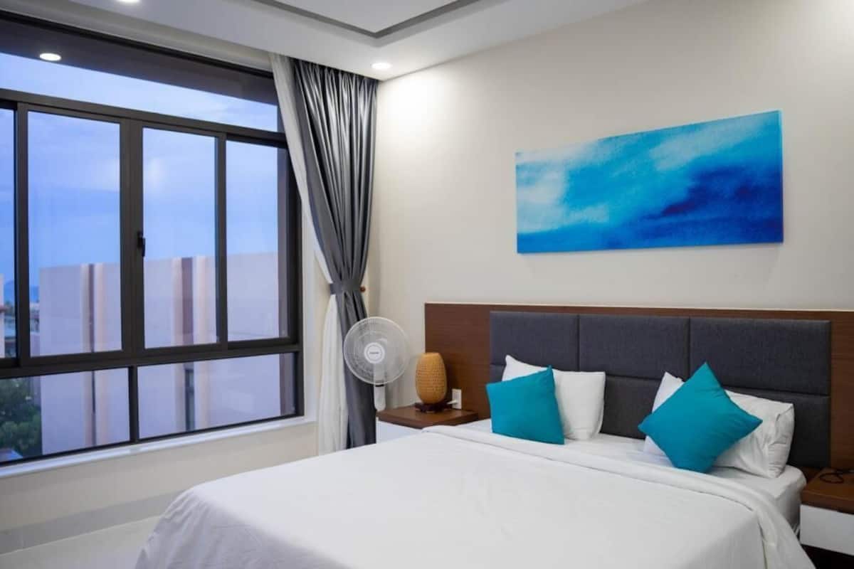 A hotel room with a large window, a bed with white bedding and blue pillows, a wall-mounted artwork, and a bedside table with a fan.