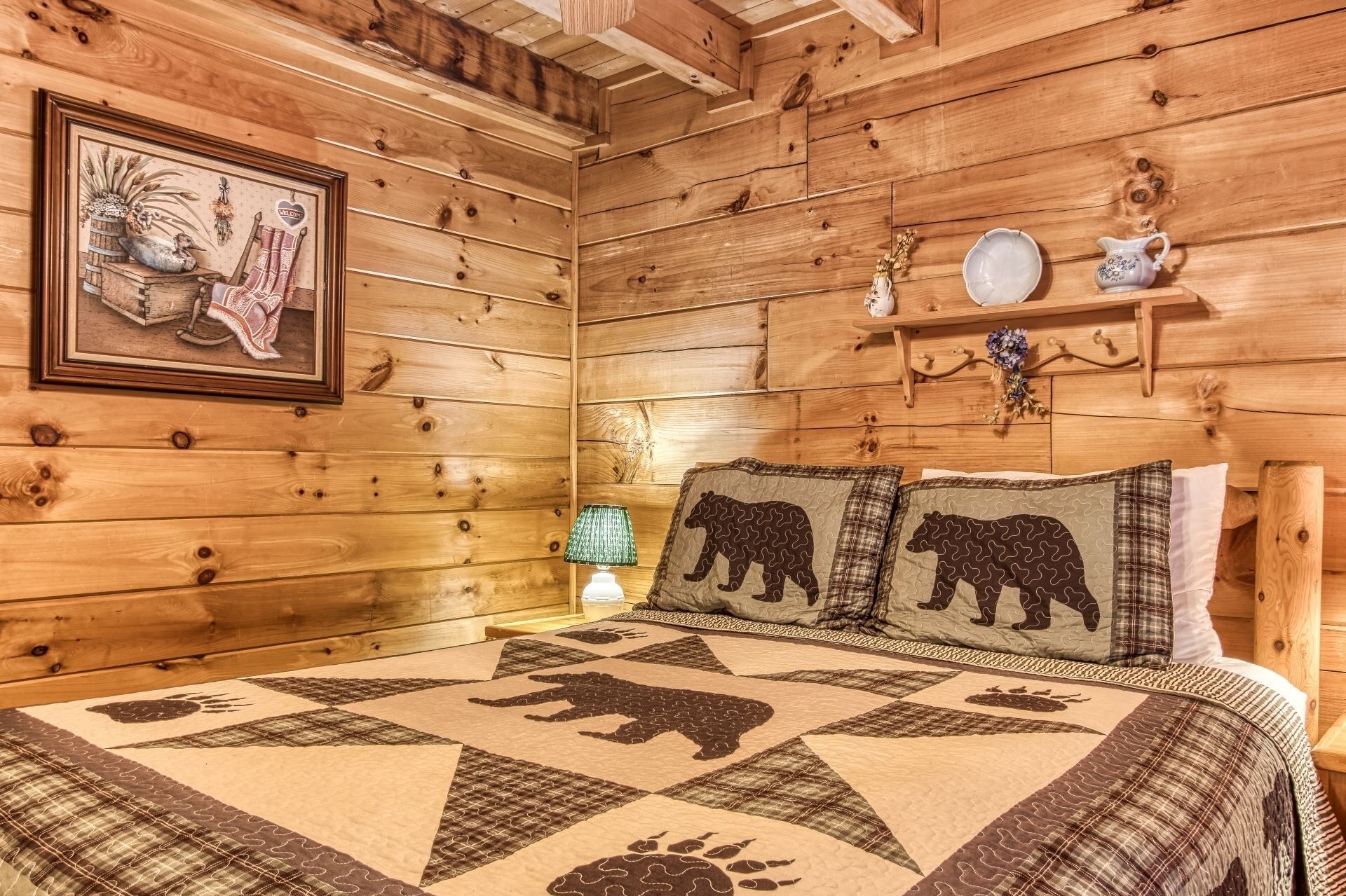 Een slaapkamer in een houten chalet met een geborduurde bedspreider met berenmotieven, een ingelijst schilderij aan de muur en een klein plankje met decoratieve objecten.