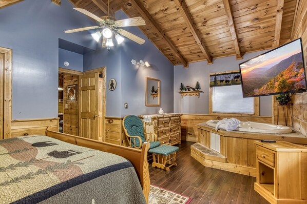 Una habitación con techo de madera, una cama con colcha, un aparador de madera, un tocador con espejo y un televisor que muestra un paisaje pintoresco.