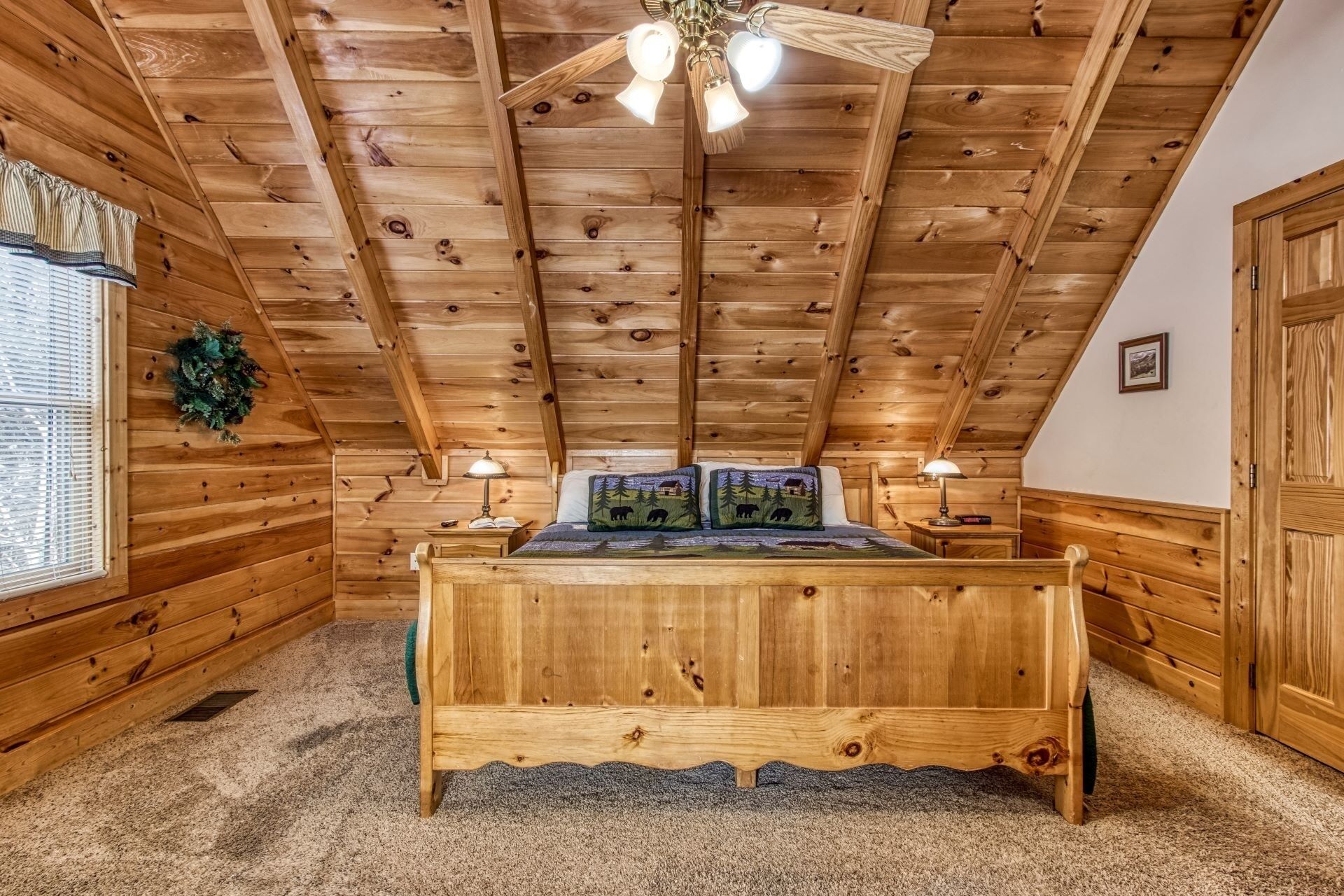 A wooden cabin bedroom with a sloped ceiling, a wooden bed, two bedside lamps, and a window with blinds.