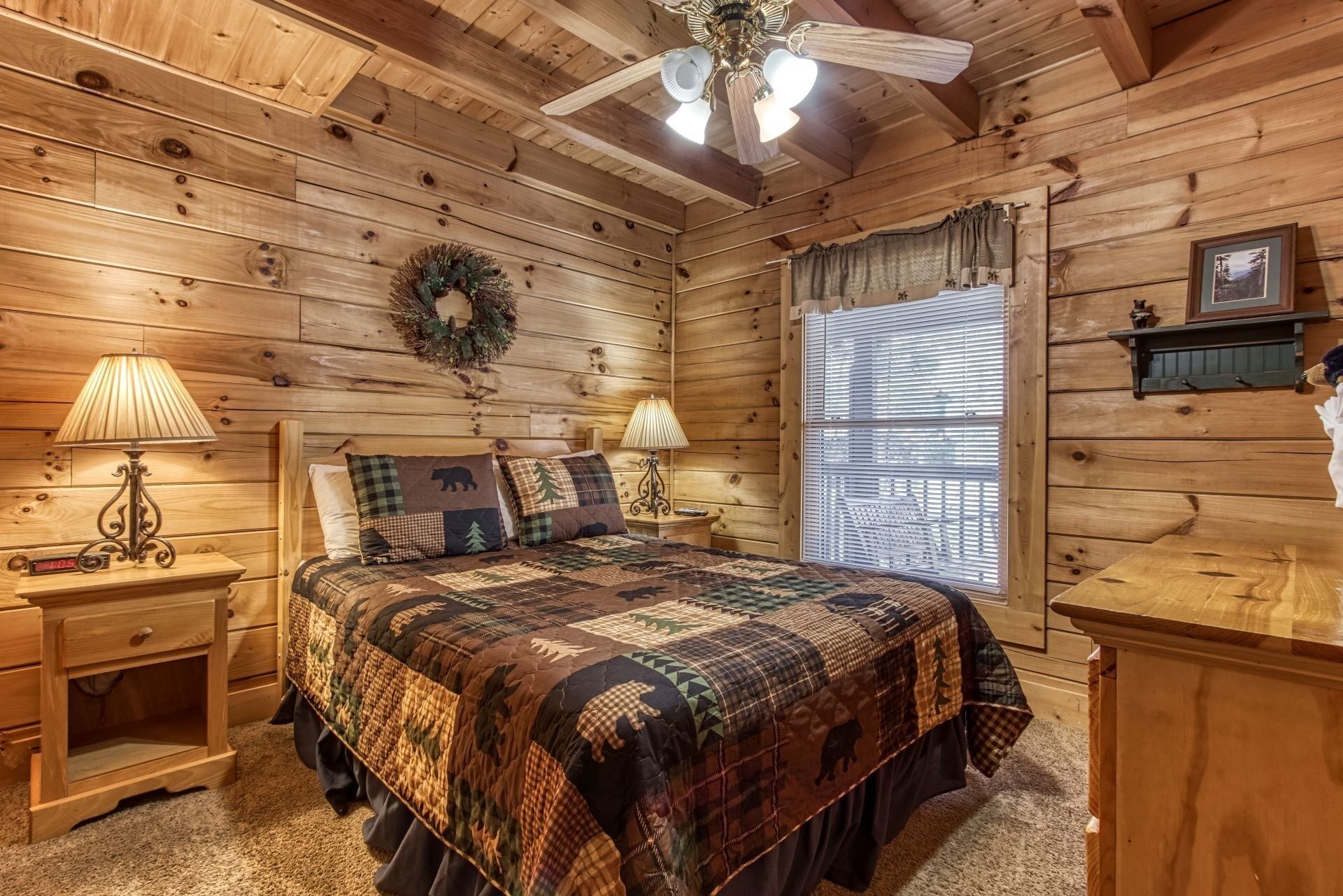 Een gezellig chalet slaapkamer met een groot bed, twee nachtkastjes met lampen, een raam met jaloezieën en een plafondventilator.