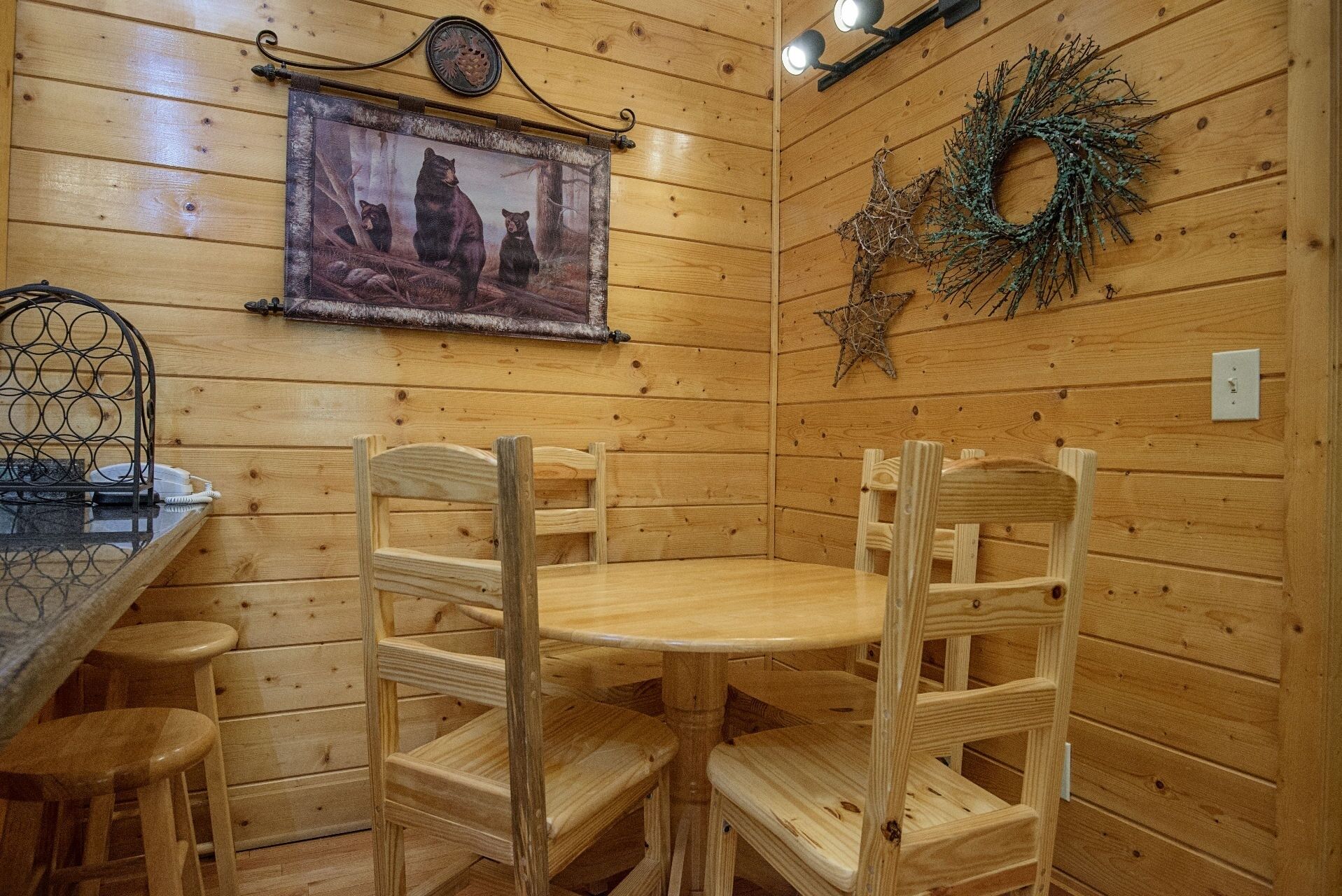 Ein Holzblockhaus-Innenraum mit einem runden Tisch und vier Holzstühlen. Ein gerahmtes Bild von Bären hängt an der Wand, und ein Kranz ist über einer Lampe angebracht.