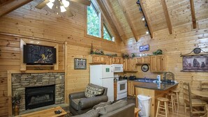 A cozy cabin interior with a stone fireplace, a leather sofa, a television, wooden cabinets, a kitchen area, and a 'Welcome' sign.