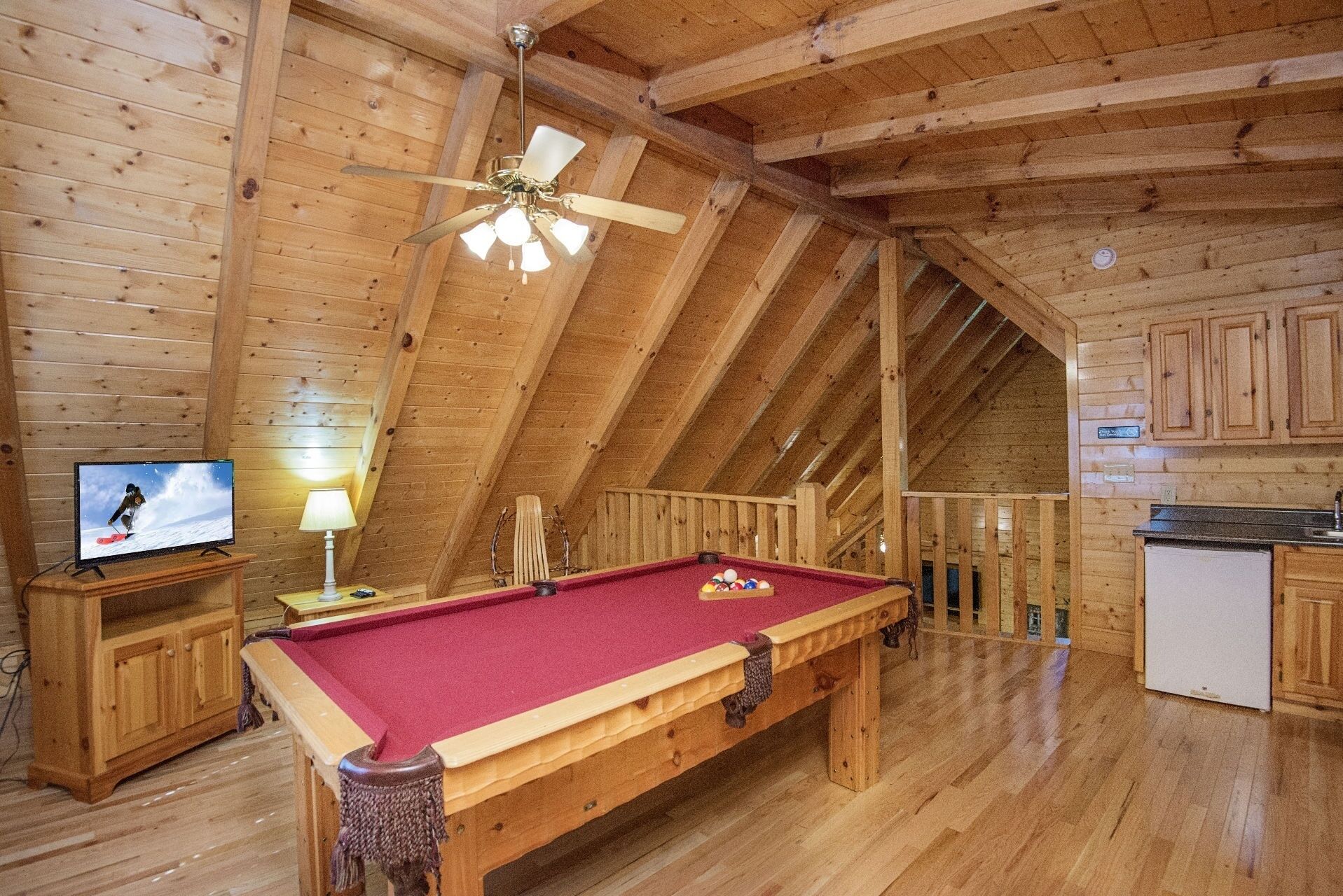 A wooden interior with a pool table, a ceiling fan, a television, and a kitchen area.