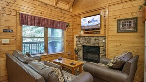 A cozy cabin interior with a stone fireplace, leather sofas, a television, and a 'Thank You For Not Smoking' sign.