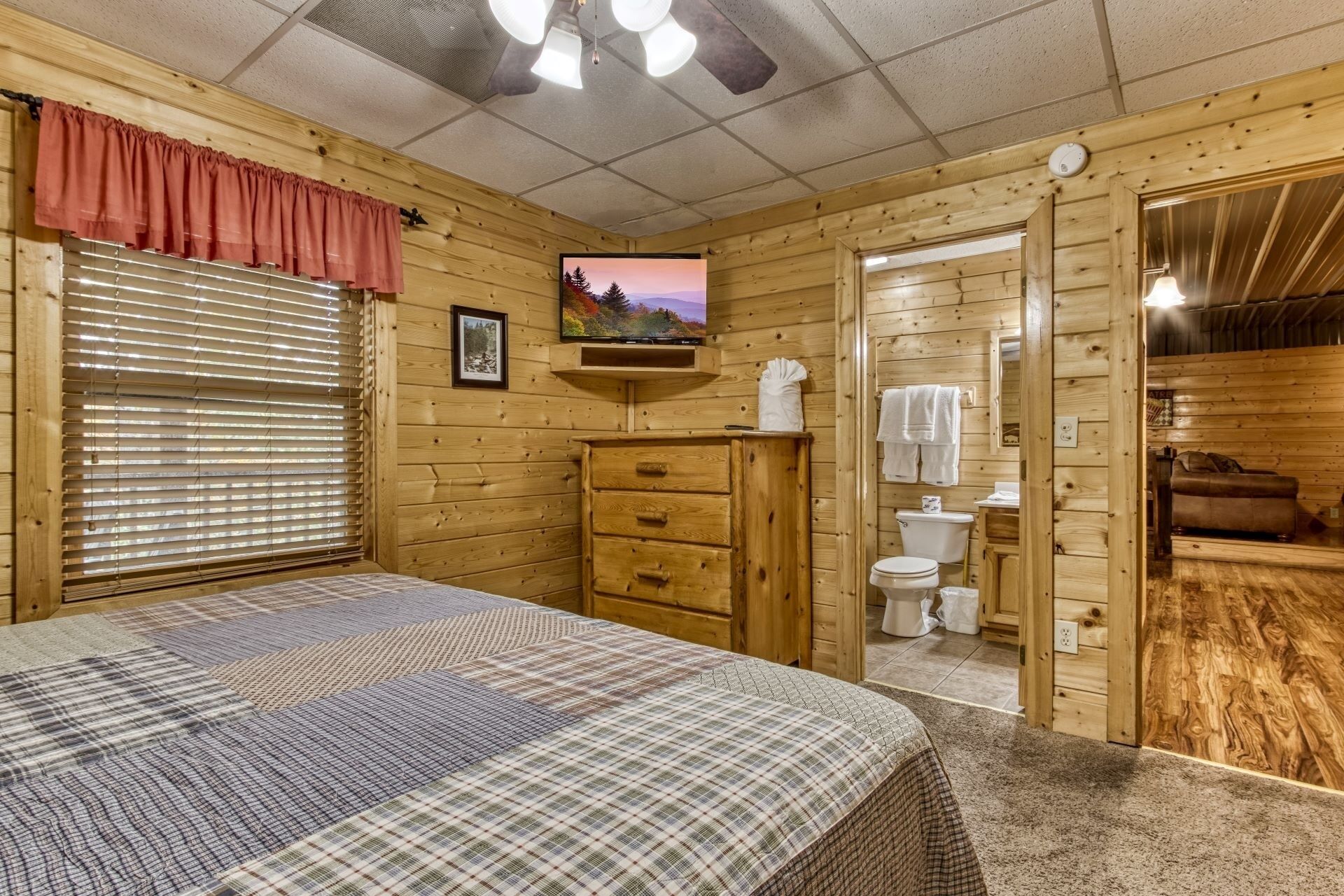 Un dormitorio de cabaña de madera con cama, cómoda, ventana con persianas, un cuadro en la pared y un baño con inodoro y lavamanos.
