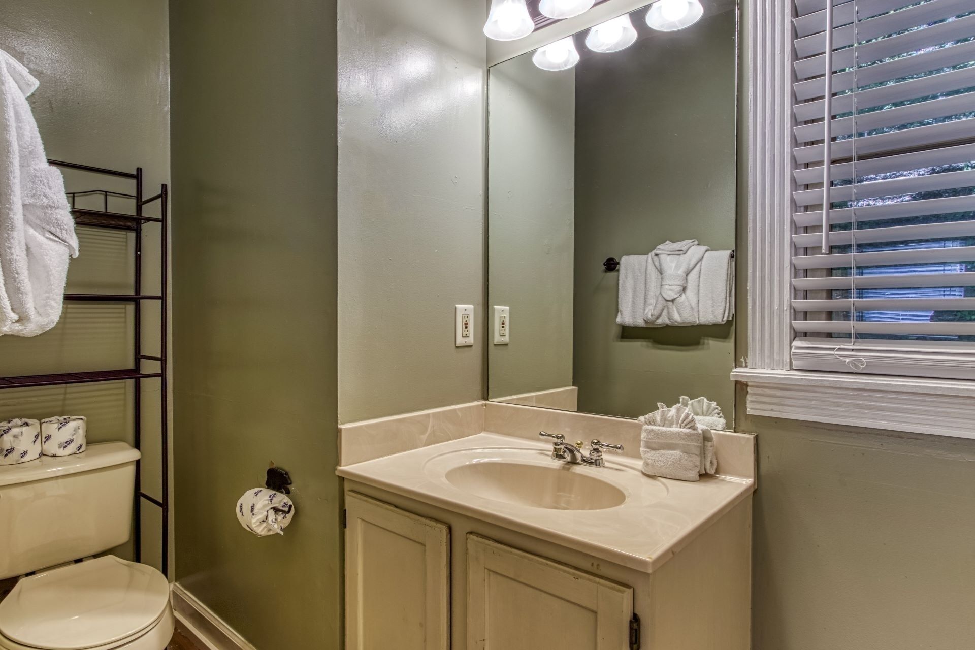A bathroom with a sink, mirror, and toilet. There are towels hanging on the wall and a window with blinds.