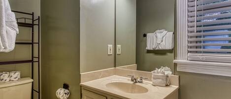 A bathroom with a sink, mirror, and toilet. There are towels hanging on the wall and a window with blinds.