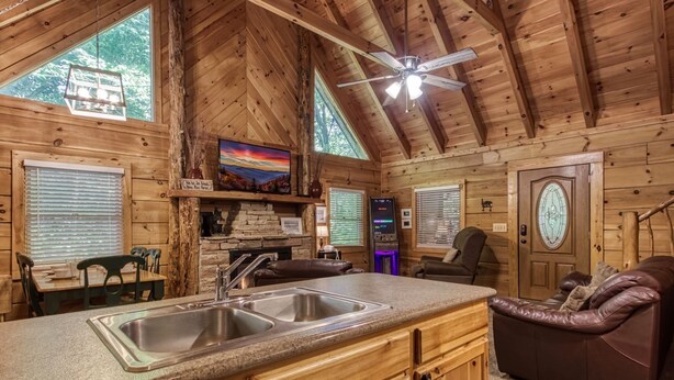 A cozy cabin interior with a kitchen area featuring a double sink, wooden cabinets, and a stone fireplace. There is a comfortable seating area with leather couches and a large window offering a view of the outdoors. - Er96 - Squirrel's Leap - Great Location! Close To Town! 2 Bedroom Cabin by RedAwning (Pigeon Forge)