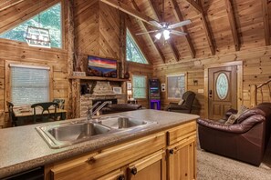 L’intérieur de ce chalet chaleureux comprend un coin cuisine équipé d’un évier à deux bacs, de meubles en bois et d’une cheminée en pierre. On y trouve également un coin salon confortable avec des canapés en cuir et une grande fenêtre offrant une vue sur l’extérieur.