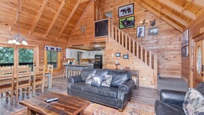 A wooden cabin interior with a black leather sofa, wooden dining table, and a staircase leading to a loft area.