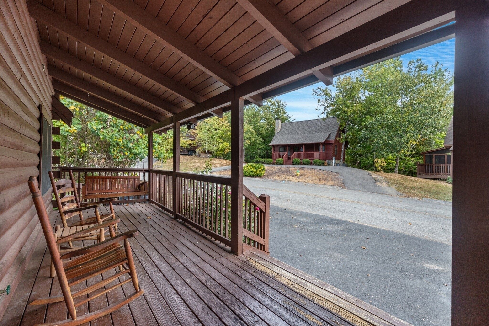 A wooden porch with rocking chairs, a covered area, and a view of a house and trees.