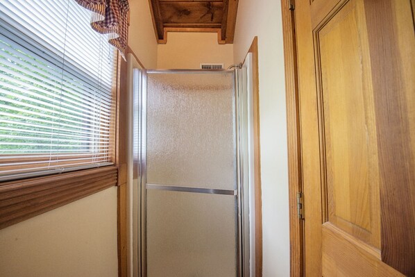 A shower with a glass door, a wooden door to the right, and a window with blinds on the left.