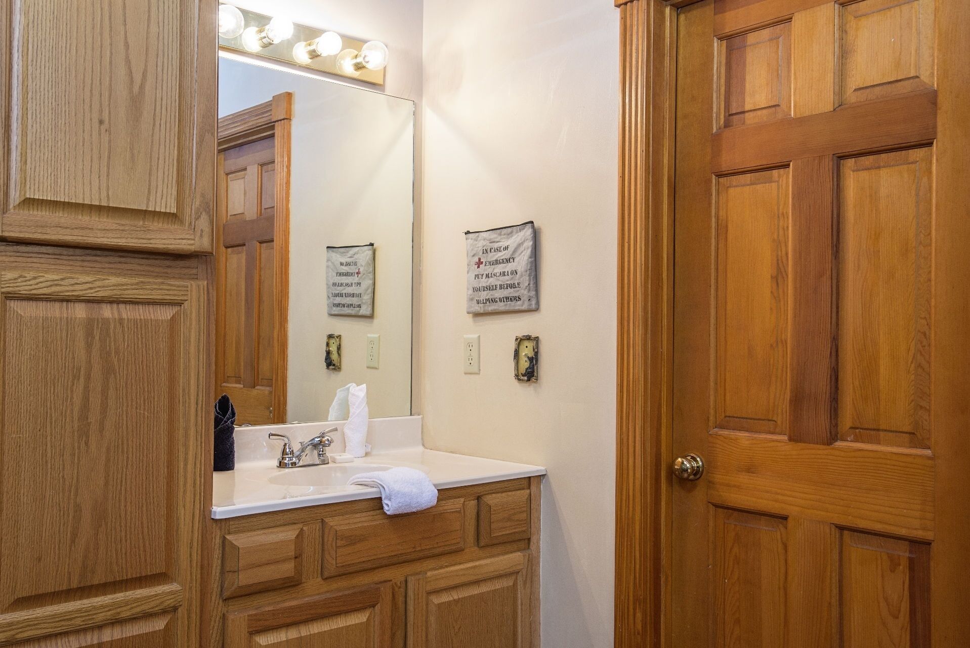 A bathroom with a wooden vanity, a mirror with lights, a towel, and a sign instructing to put on a mask in case of emergency.