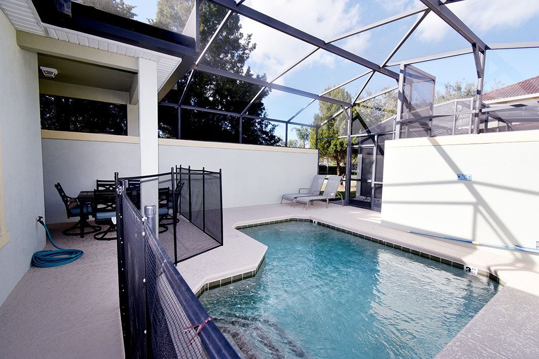 A swimming pool with a glass enclosure, a patio area with chairs, and a view of the surrounding landscape.