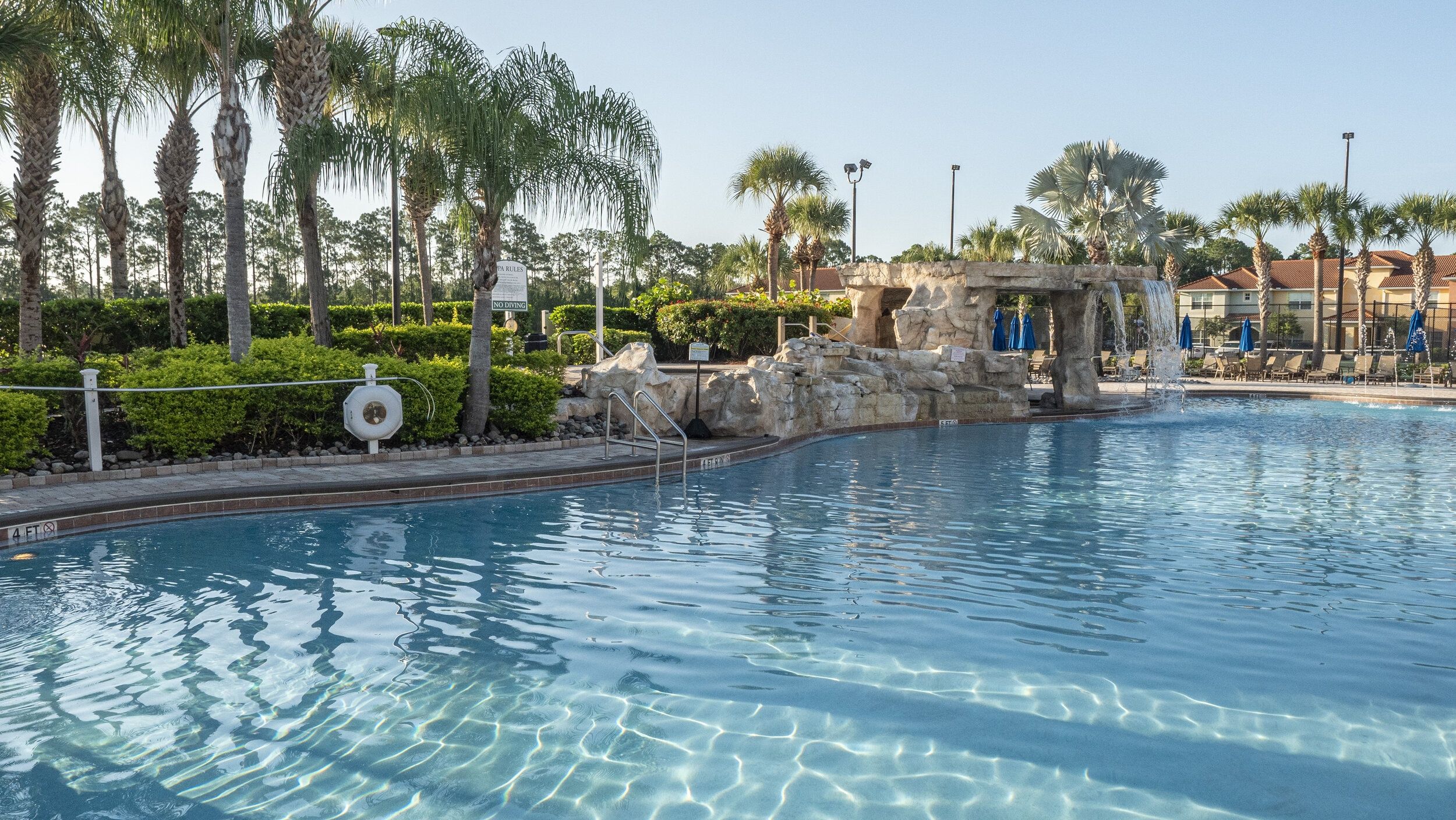 A clear swimming pool with a rock feature, surrounded by palm trees and a building in the background.