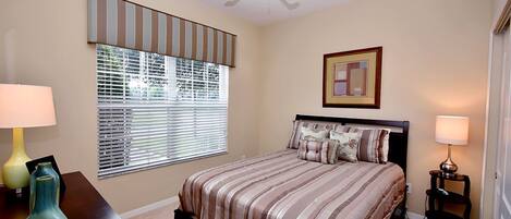 A bedroom with a bed, a ceiling fan, a window with blinds, a bedside table with a lamp, and a framed picture on the wall.