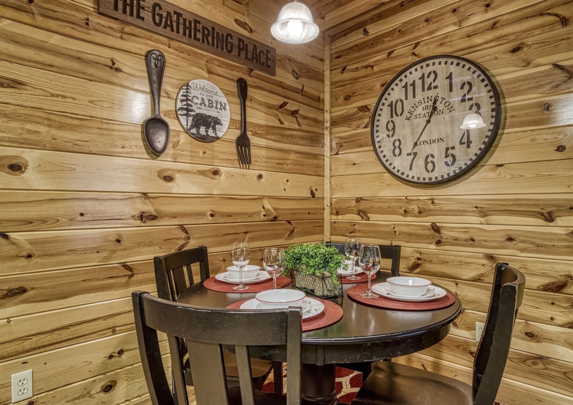 A wooden cabin interior with a round table set for dining, a wall clock, and a sign reading "The Gathering Place".
