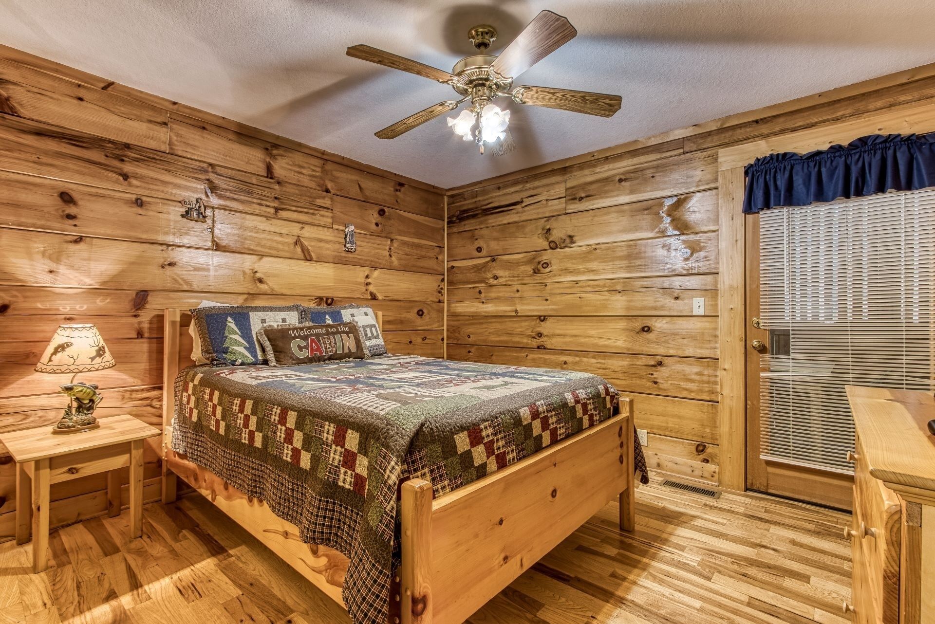 Un dormitorio tipo cabaña con cama de madera, mesita de noche con lámpara, ventilador de techo y ventana con persianas.