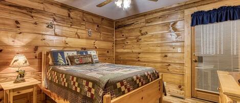 Un dormitorio tipo cabaña con cama de madera, mesita de noche con lámpara, ventilador de techo y ventana con persianas.