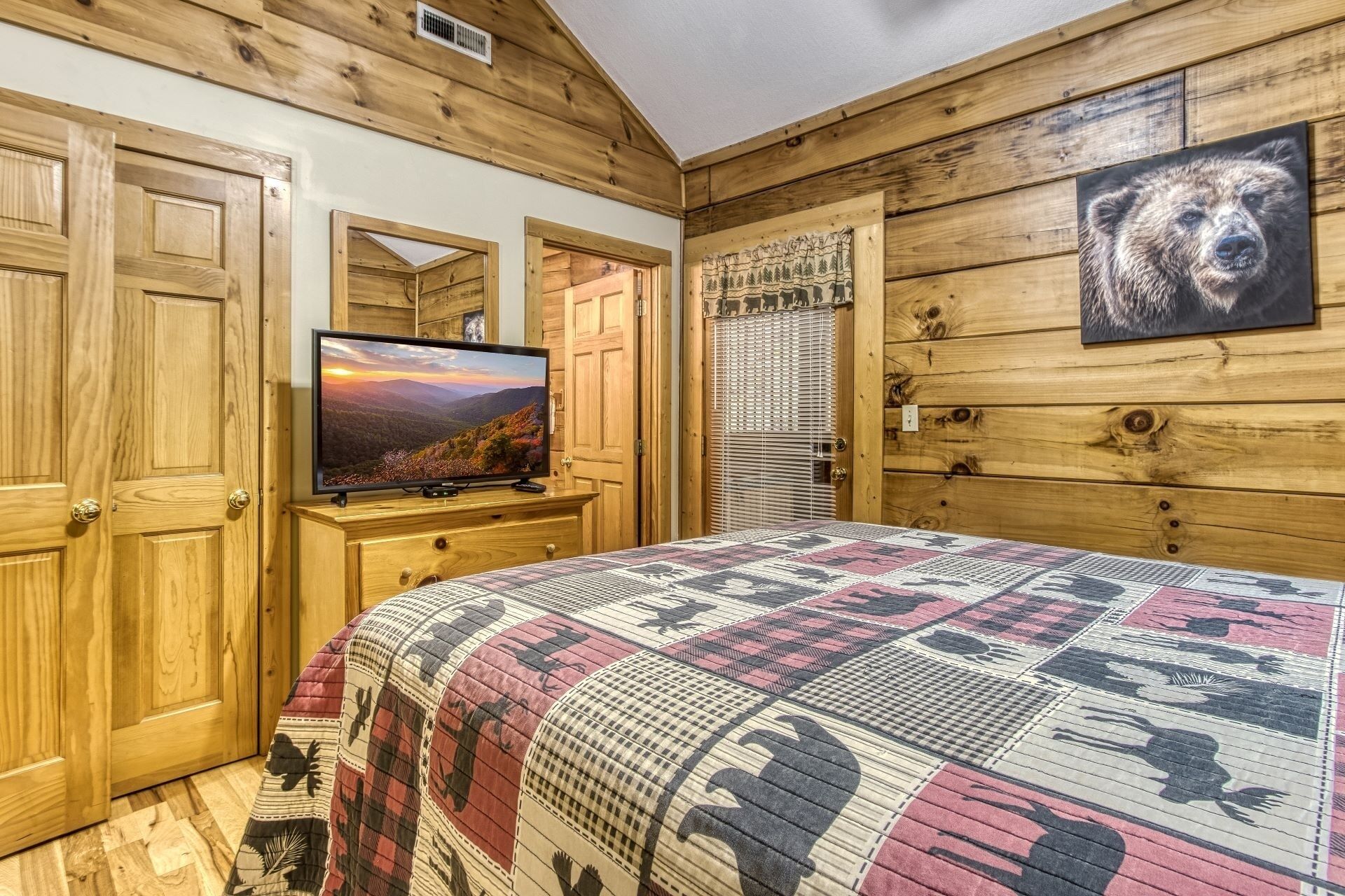 A bedroom with wooden walls, a bed with a patterned quilt, a television on a wooden dresser, and a bear painting on the wall.