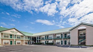 Un motel con techo verde y varias habitaciones, un estacionamiento y un cielo azul despejado con nubes.