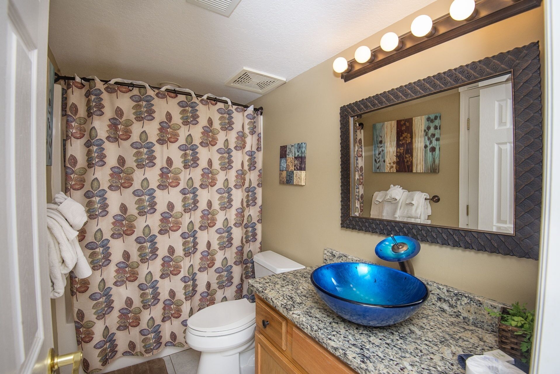 Un baño con inodoro, un lavabo de color azul, cortina de ducha con diseño de hojas, espejo con marco decorativo y una lámpara empotrada en la pared.