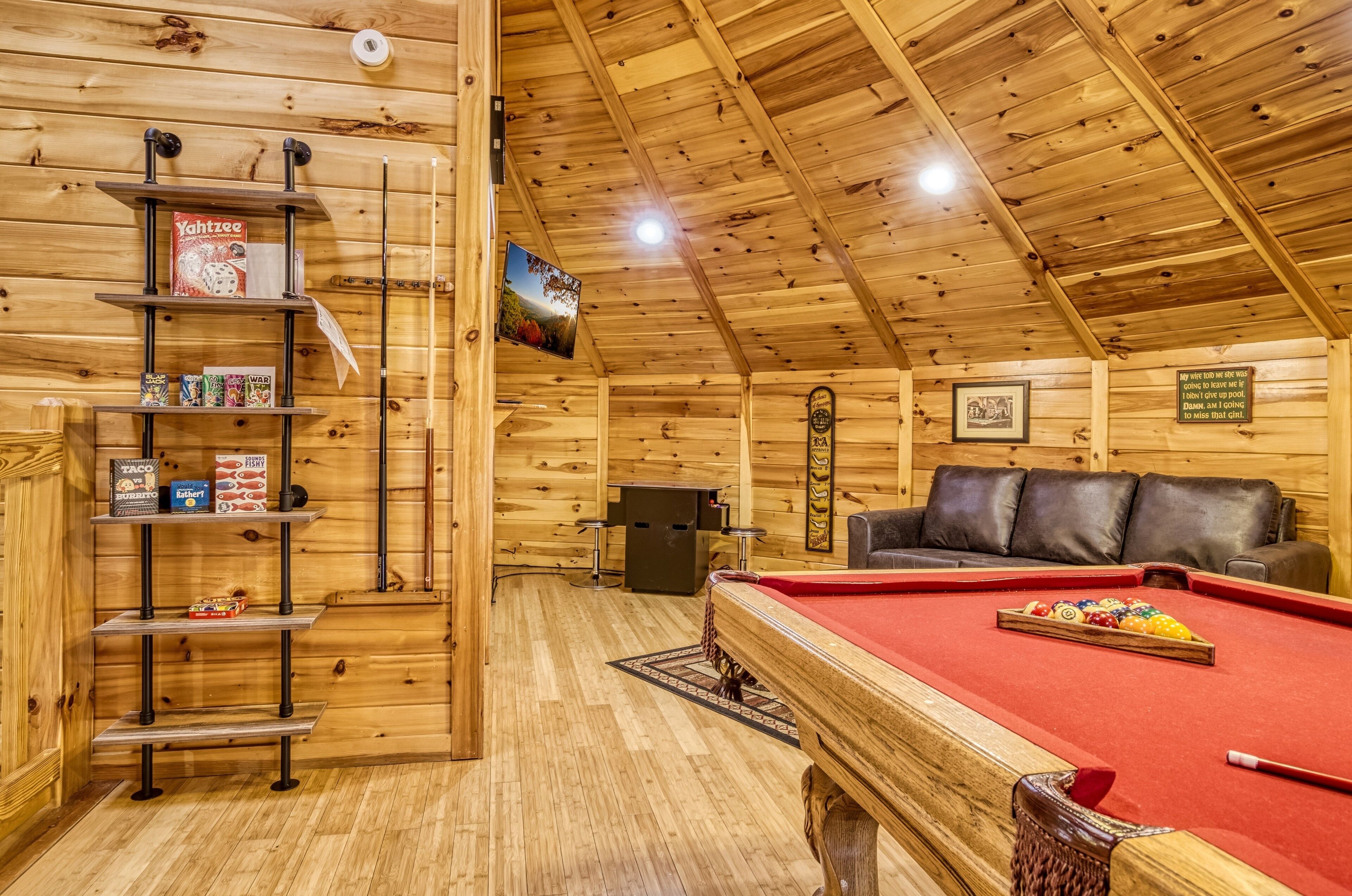Une pièce en bois avec une table de billard, des étagères remplies de collations, un canapé en cuir et un téléviseur fixé au mur.