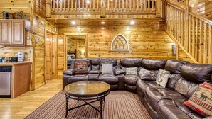 A log cabin interior with a leather sofa, wooden walls, a staircase, and a window with a decorative arch. - Declan's View - Cozy 1 bedroom with Game room and Great Mountain Views! 1 Cabin by RedAwning (Sevierville)