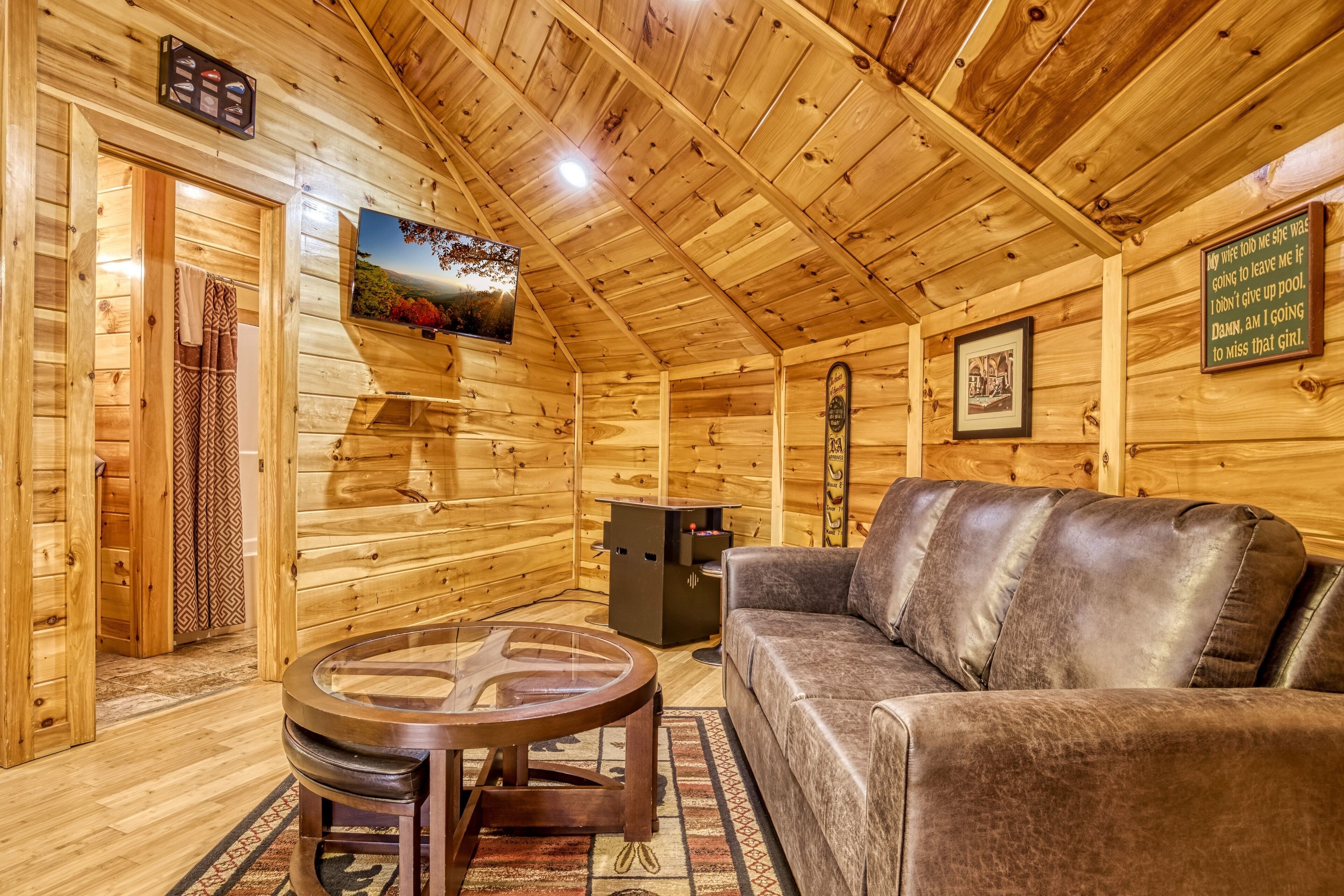 A wooden cabin interior with a leather sofa, a round coffee table, and a television mounted on the wall.