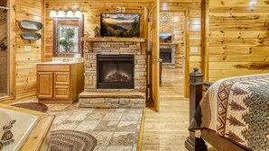 A log cabin interior with a stone fireplace, wooden walls, a large mirror, and a bed with a patterned blanket. - Declan's View - Cozy 1 bedroom with Game room and Great Mountain Views! 1 Cabin by RedAwning (Sevierville)